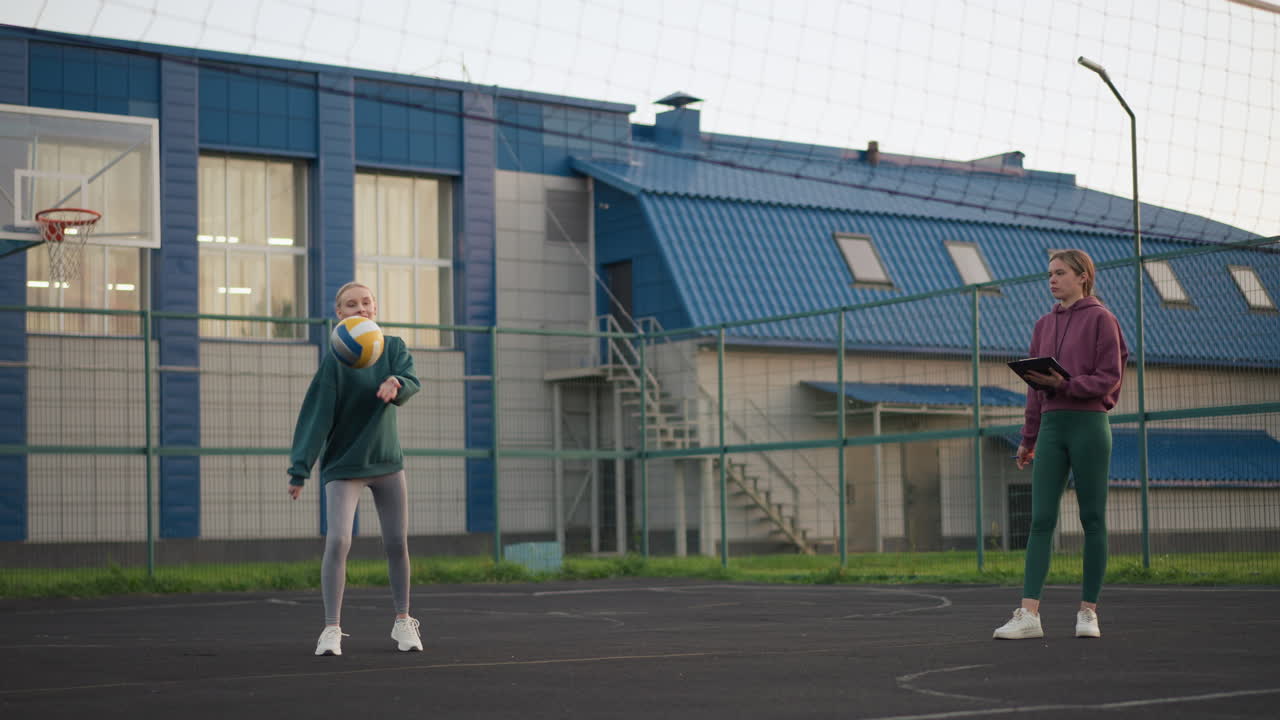 начинающий волейболист наблюдается тренером во время подачи с книгой результатов в руке и свистком во рту, наблюдение за техникой на открытом корте с сосредоточенным вниманием