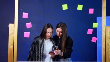 two ladies stand behind the glass board with stickers. women look focused on the phone watching video.