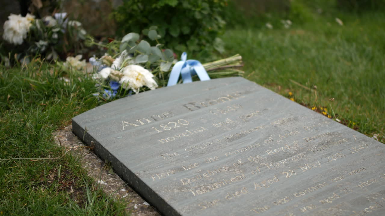 A gravestone marked "Anne Brontë" is surrounded by white and blue flowers laid on grass. Historical marker, cemetery scene, and literary memorial are prominently featured