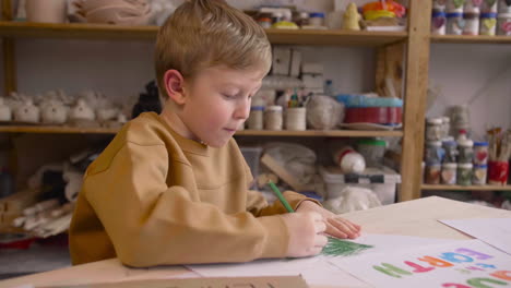 Niño-Rubio-Dibujando-Con-Lápiz-Verde-Sentado-En-Una-Mesa-En-Un-Taller-De-Artesanía-Donde-Hay-Carteles-Con-Citas-Ambientales