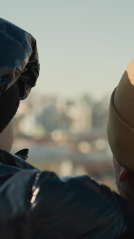 un padre que lleva a su hijo mira hacia un paisaje urbano, mientras que el hijo mira a la cámara antes de darse la vuelta, ambos están vestidos con chaquetas y gorras, con el paisaje urbano borroso