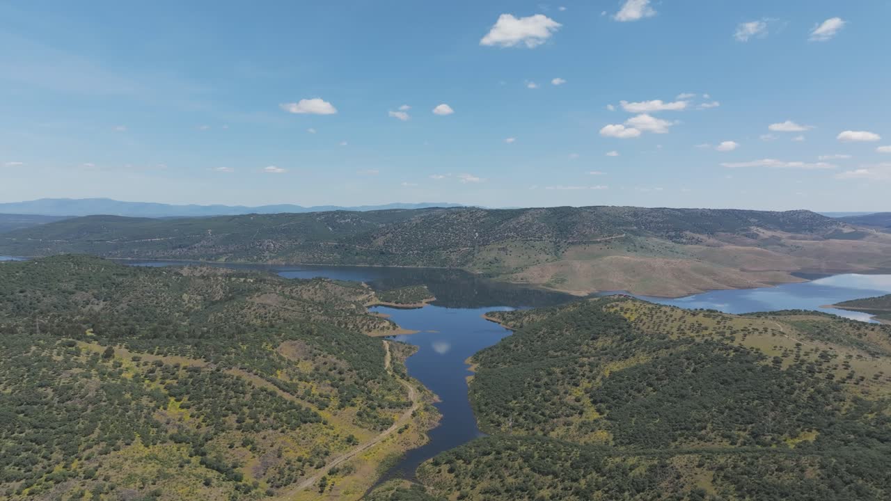 Drone shot sweeping over Cíjara Reservoir in Badajoz, Spain, with calm blue waters, clouds reflecting, green hills and dry grass under a clear sky. Ideal spot for hiking, fishing and outdoor life