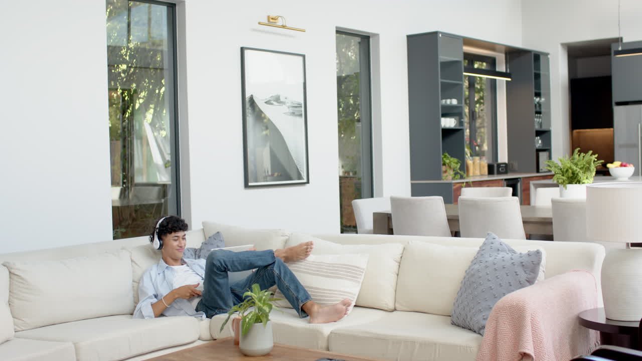 Relaxing on sofa, teenage boy with headphones using tablet in modern living room