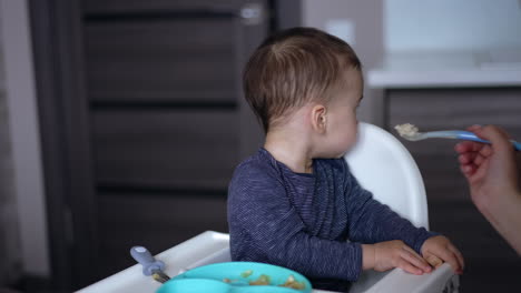 cute baby sitting in the feeding table and turning. mom shoves the spoon to a kid's mouth. blurred backdrop.