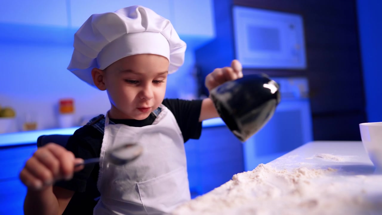 Bambino piccolo con cappello da chef che tiene un cucchiaio. La farina si sparge sul bancone bianco mentre il bambino lavora in una cucina illuminata di blu