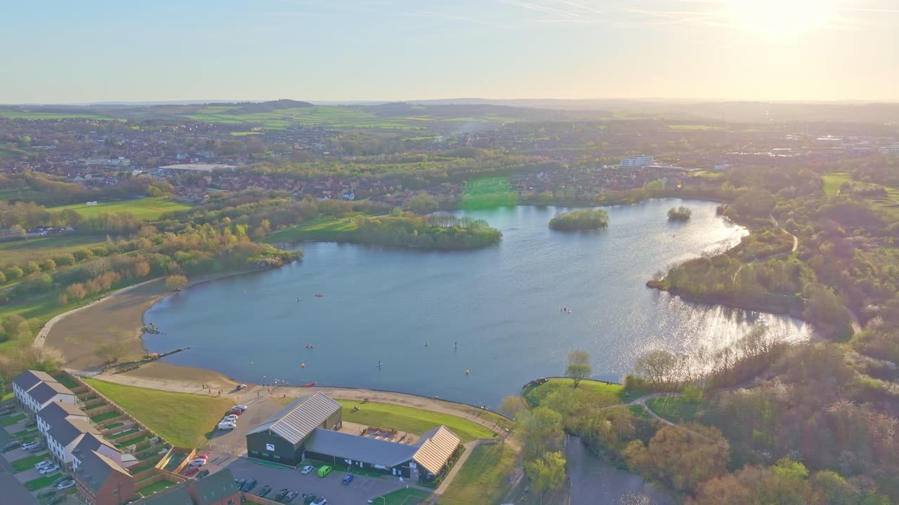 Expansive drone view of Manvers Lake in Rotherham, South Yorkshire, at golden hour showing reflective water, activity centre with solar panels, green areas and distant townscape, captured in real time