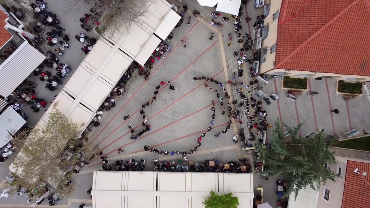 Vista aerea di una danza popolare tradizionale che si snoda a spirale attraverso la piazza del villaggio, Deskati, Grecia. I ballerini formano una fila sinuosa tra edifici con tetti rossi