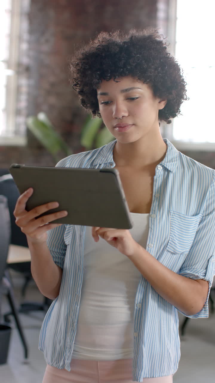 Vertical video of portrait of happy biracial casual businesswoman using tablet in slow motion