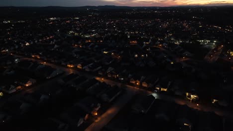 Night-aerial-view-of-a-suburban-neighborhood,-illuminated-by-streetlights-and-house-lights,-with-a-backdrop-of-distant-city-lights-and-a-colorful-sunset