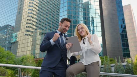 euphoric businessman and businesswoman with tablet pc in business district with skyscrapers celebrating successful deal or trade, online lottery win or auction bid, excited of reading great news
