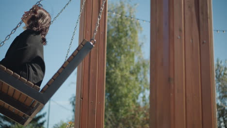 vista lateral de cerca de una mujer sentada en un columpio de madera, el cabello soplando suavemente en el viento, rodeada de exuberante vegetación en un tranquilo parque