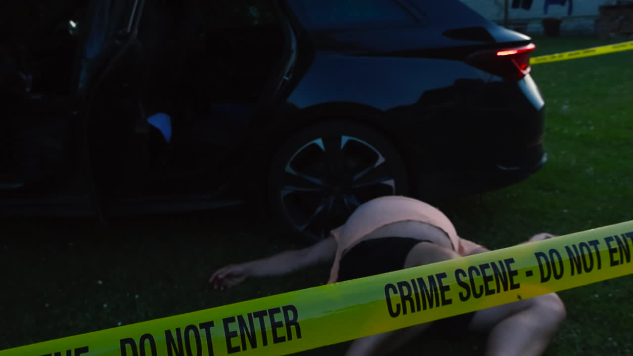 Police investigation line forensics yellow colored Crime Scene tape hanging and pulled in front of a black crashed car where a fat victim is lying on the ground during evening, car lights are blinking
