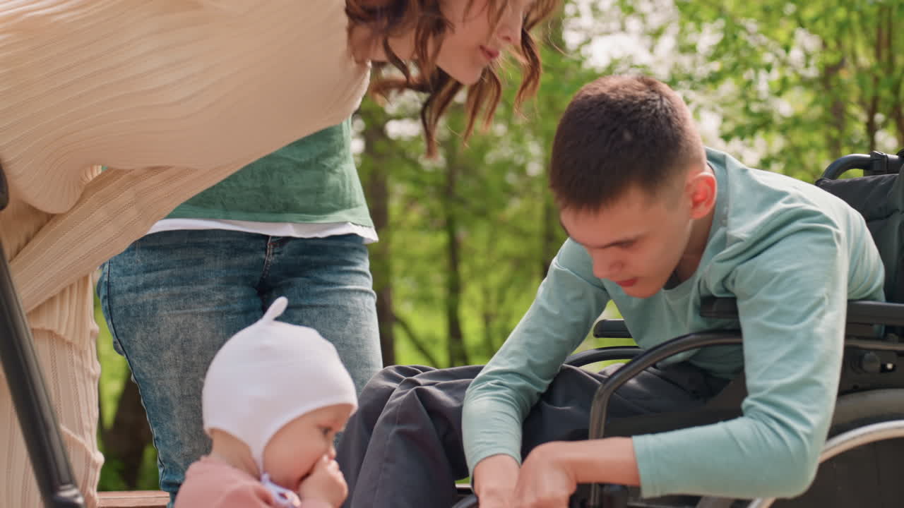 Caregiver Assisting Young Man In Wheelchair, Park Scene Showing Close Attention, Mother And Grandmother Nearby, Baby In Stroller, Tender Interaction And Supportive Bonding, Calm Greenery Background