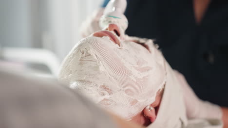 cosmetologist gloved hand guides vibrating electric facial roller in circular strokes over thick white cream mask on reclined spa client under bright lamp, capturing mask texture