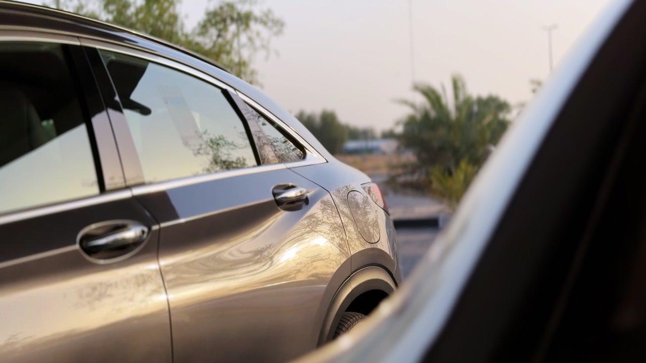 side profile of a modern dark metallic sedan parked outdoors during golden hour. The setting sun reflects off the vehicle's polished bodywork with soft light hitting the car exterior.