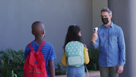 Diverse-male-teacher-measuring-temperature-of-schoolchildren,-all-wearing-face-masks