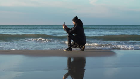 Man-squats-taking-photo-of-beach-low-tide-wet-sand,-with-waves-background