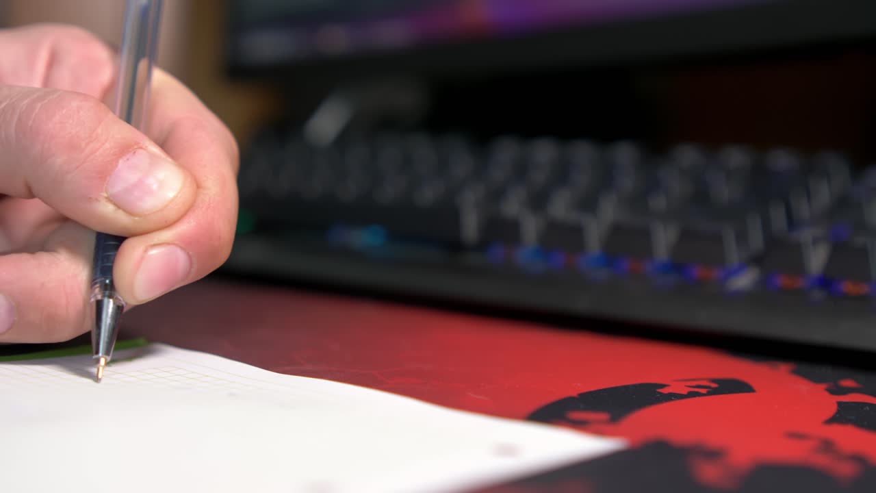 Una mano scrive su carta sopra una superficie con motivo, con l'illuminazione dinamica di una tastiera sullo sfondo. L'attenzione è sul processo di scrittura e sull'illuminazione colorata.