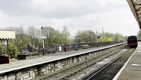 A-slow-motion-video-of-an-old-fashoned-steam-train-arrives-from-the-distance-and-begins-to-slow-as-it-approaches-the-station-platform,-on-a-cloudy-day-in-the-United-Kingdom