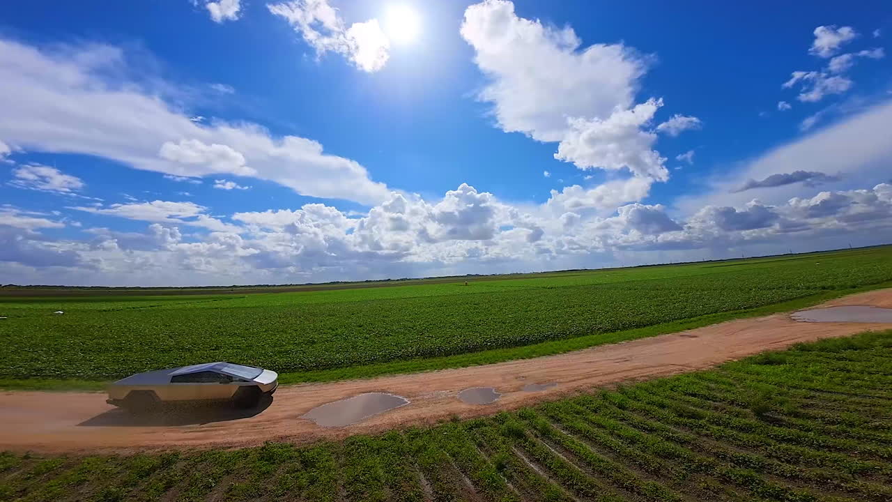 Moving Tesla Cybertruck in the green fields. Fast electric car rides by the puddles on the ground road. Beautiful azure sky with fluffy clouds at backdrop.