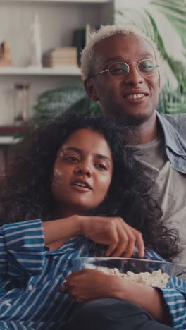 young happy multiethnic couple eating popcorn watching movie on tv sits on sofa
