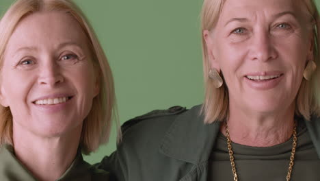 close up view of two blonde mature women and blonde senior woman smiling and hugging, wearing green tones clothes and posing on green background