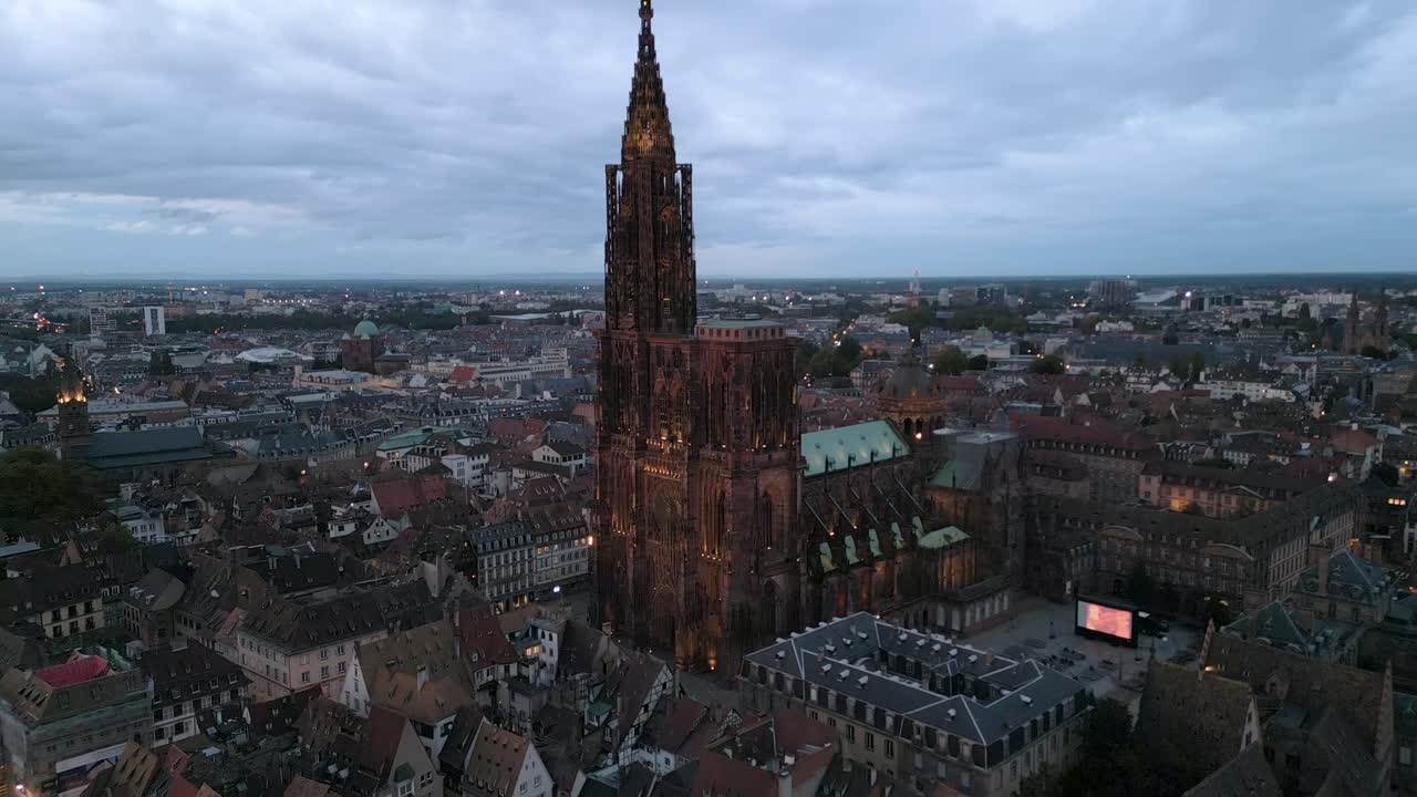Drammatica ripresa aerea ravvicinata al crepuscolo della Cattedrale di Strasburgo che svetta sui tetti medievali della Città Vecchia di Strasburgo. Le luci della città iniziano a scintillare mentre il crepuscolo cala su questo storico paesaggio urbano francese.
