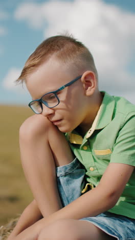 un niño pequeño se sienta en una bola de heno con la cabeza inclinada, perdido en sus pensamientos, rodeado de un vasto campo abierto bajo un cielo azul brillante, capturando un momento tranquilo y pacífico en el campo en un cálido día soleado.