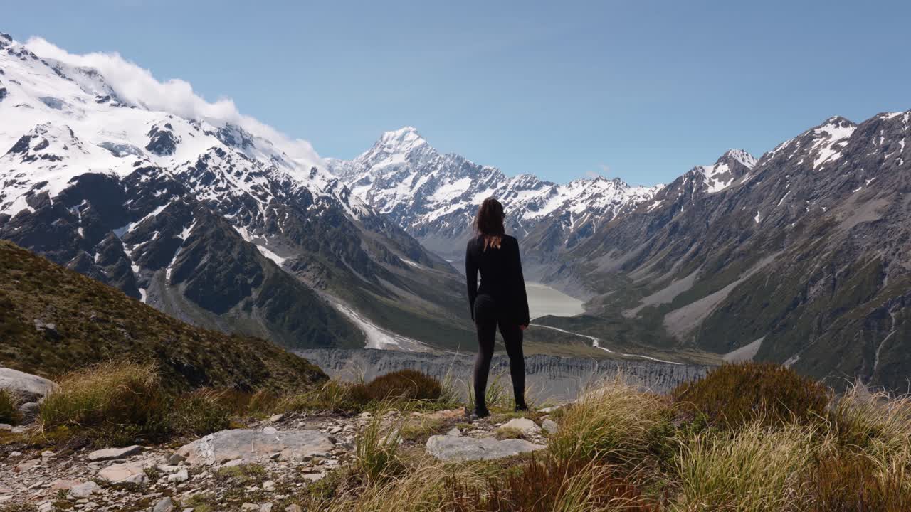 Young caucasian woman walking and looking around at Sealy Tarns viewpoint on a sunny summer day wih Hooker Lake, and snowy mountain peaks in background in Mount Cook National Park, New Zealand.