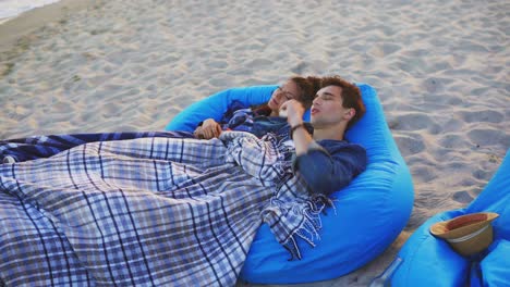 pareja acostada en sillas de descanso en la playa cubierta con una manta. hombre fumando cigarrillo electrónico. vaper. filmado en 4k