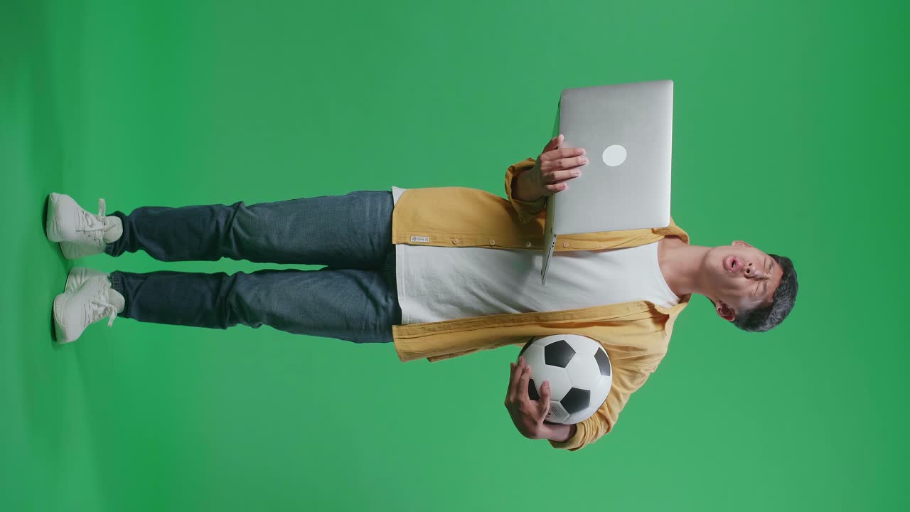 cuerpo lleno de niño asiático con una pelota se arrepiente animando al fútbol en una computadora portátil en el estudio de pantalla verde