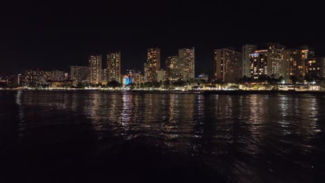 30-Fps-Nächtliche-Drohnenaufnahmen-Von-Waikiki-Hawaii,-Das-In-Den-Streifen-Mit-Skyline-Und-Verkehr-In-Honolulu-Fliegt,-Mit-Wellen-Und-Stadtlichtern-Und-Palmen