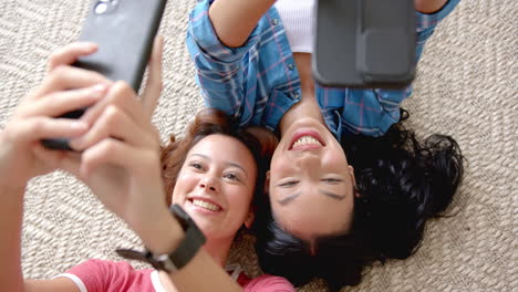 two young biracial female friends are lying on the floor, taking a selfie with their phones