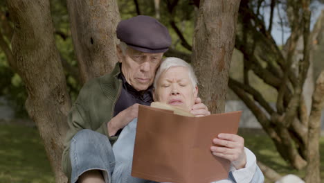 pareja mayor leyendo un libro mientras se sienta bajo un árbol y pasa tiempo en el parque en un cálido día de otoño