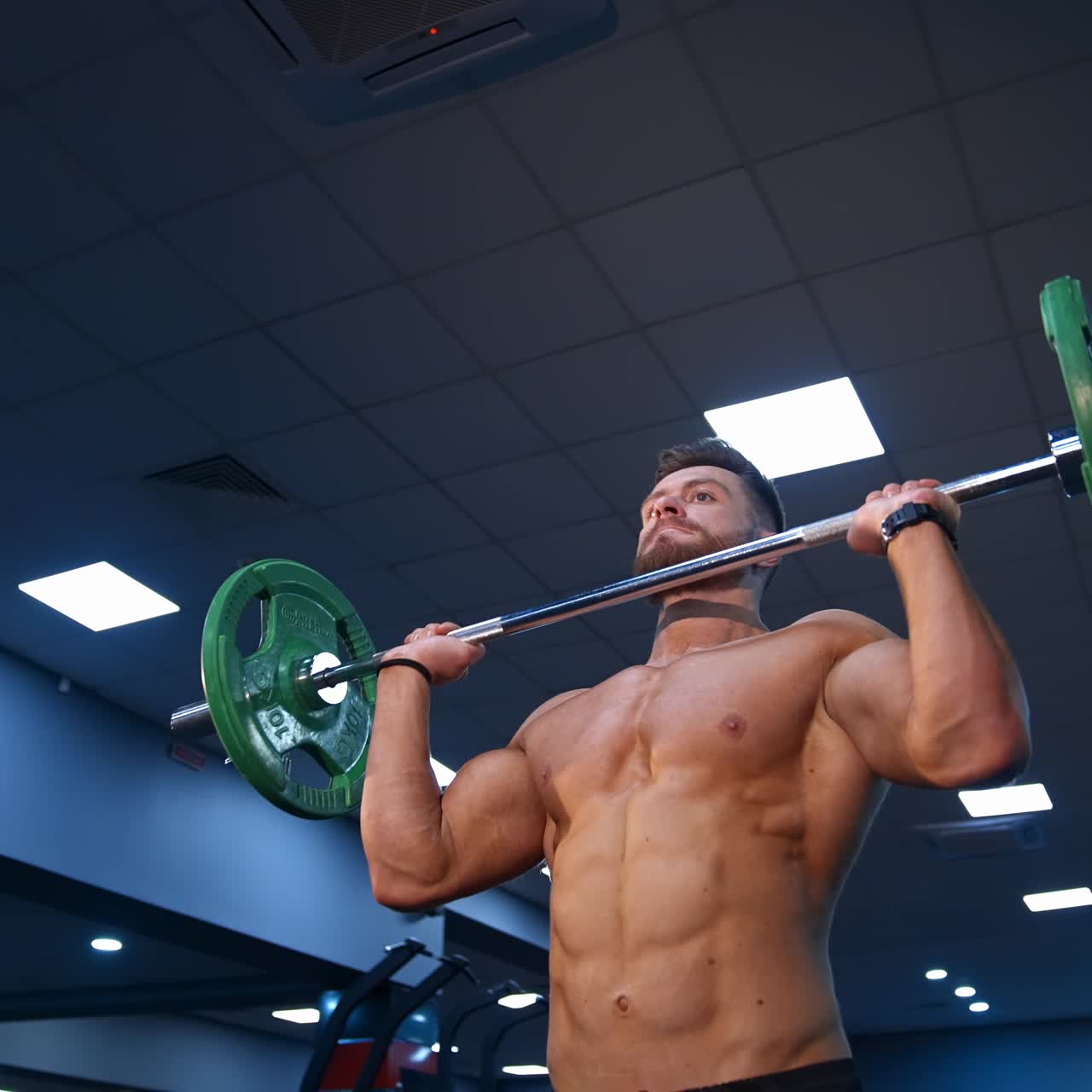 Handsome male bodybuilder lifts barbel over head. Selective focus on shirtless athlete. Slow motion