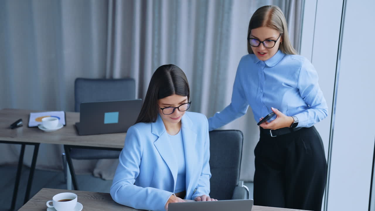 Ladies working together in office discussing job issues. Female co-workers look at laptop and taking notes. Office backdrop.