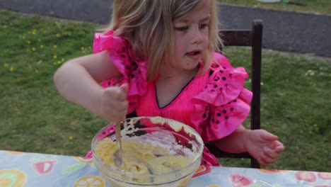 mid shot of enthusiastic young girl in pink princess dress mixing a bowl of cake mix on a patio table in daytime