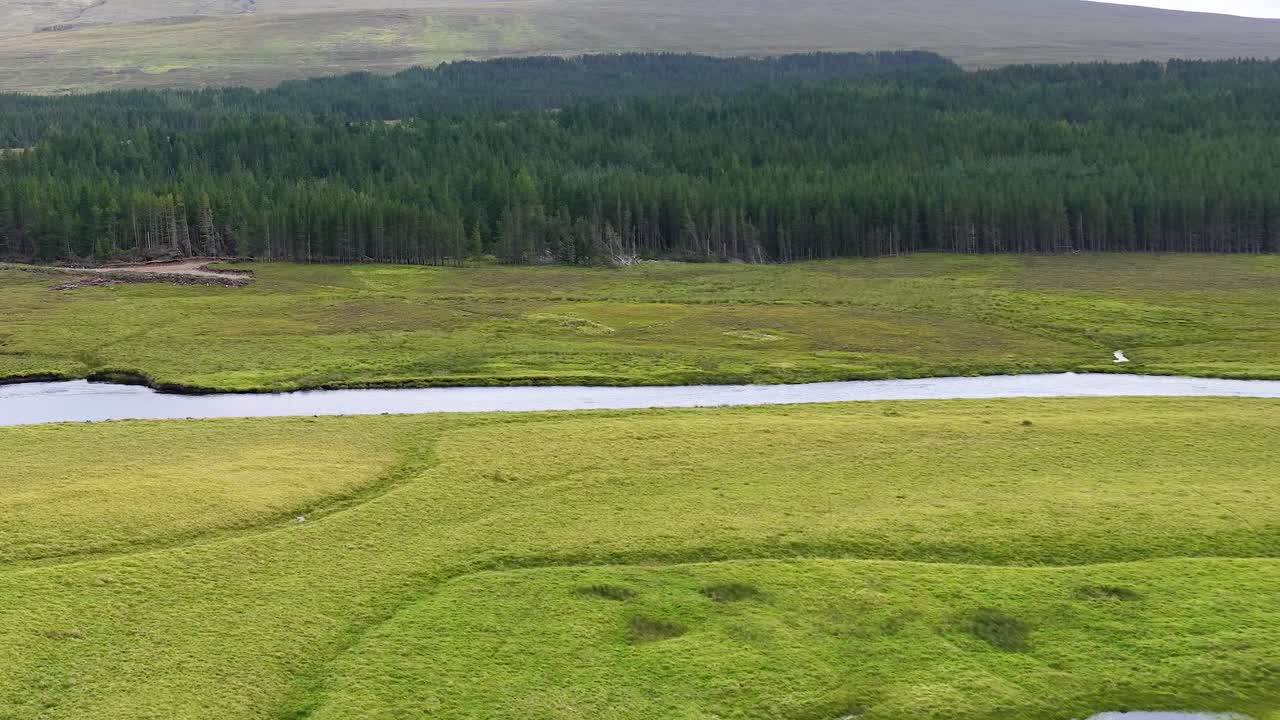 Wide shot pans over lush river, meadows, forest, and hills under soft natural daylight