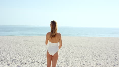 Hermosa-Mujer-Atándose-El-Cabello-En-La-Playa-Usando-Un-Elegante-Traje-De-Baño-De-Una-Pieza-De-Diseñador-Tiene-Una-Figura-Atlética-Delgada