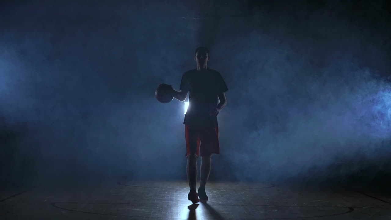 el jugador de baloncesto va directamente a la cámara en una habitación oscura con una luz de fondo en el humo mirando a la cámara en cámara lenta. steadicam