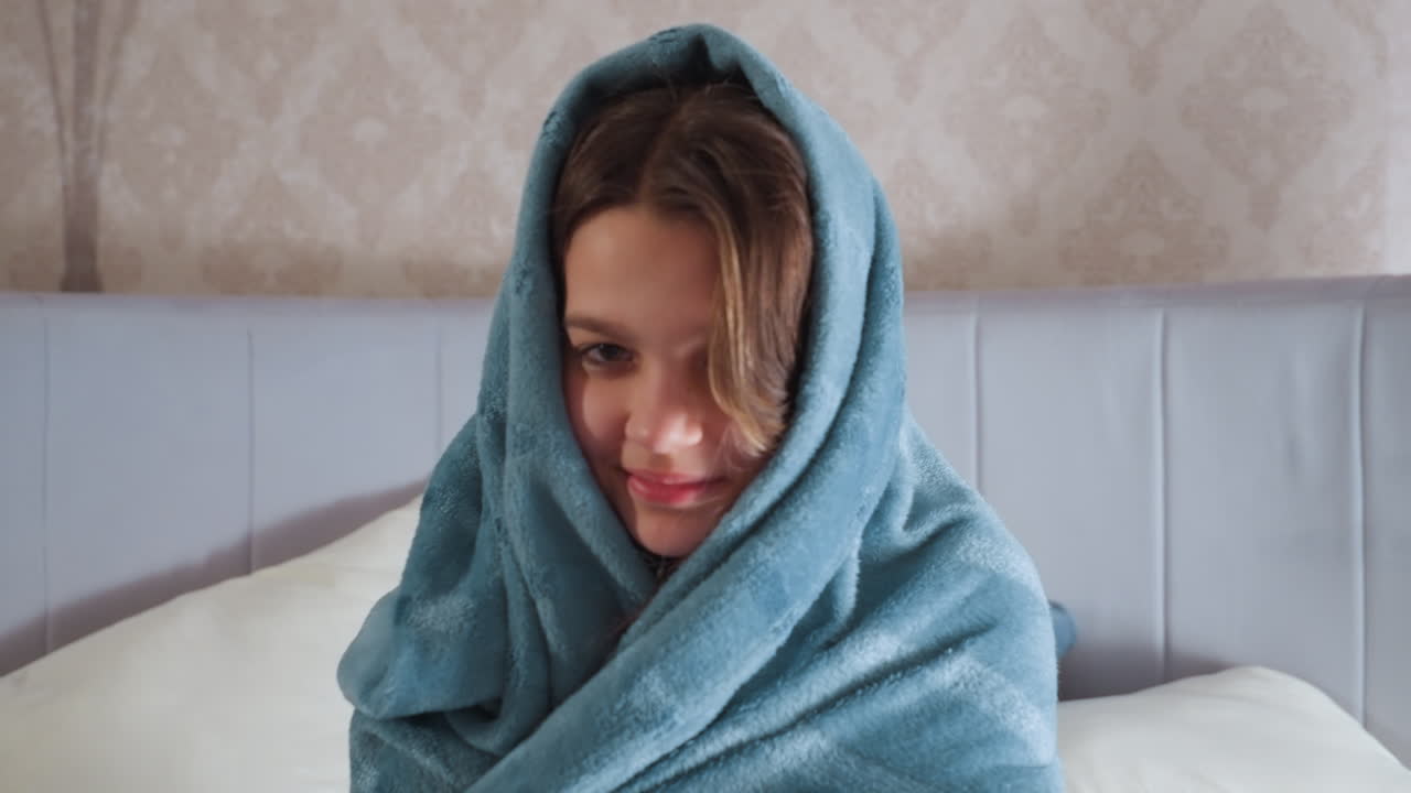 Young girl wrapped in soft blue blanket with hood covering head sits comfortably on bed, smiling softly in warmly lit bedroom with pillows and armchair