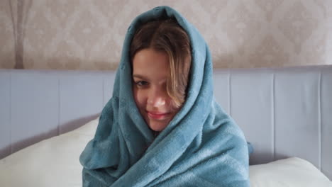 young girl wrapped in soft blue blanket with hood covering head sits comfortably on bed, smiling softly in warmly lit bedroom with pillows and armchair