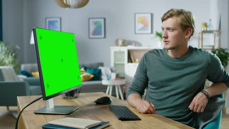 handsome young man works on a green mock-up screen personal computer while sitting at his desk in the cozy apartment.