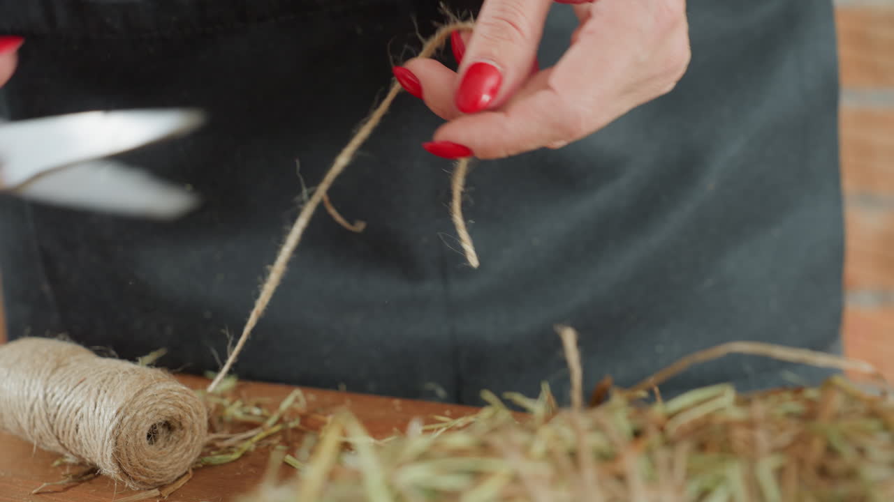 Close up of female hands with red nail polish tying twine around straw wreath on wooden table, focusing on handmade crafting, rustic decoration, and artisan detail in floral arrangement preparation