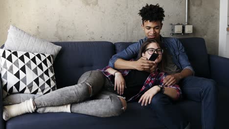 cute young couple on the couch, attractive caucasian girl lying on her handsome mulatto boyfriend's laps. he is trying to find a perfect tv chanel, with the remote in his hand.