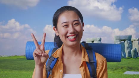 close up of asian female hiker with mountaineering backpack smiling and showing peace gesture while traveling in stonehenge