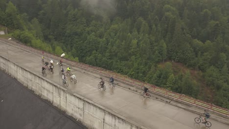 Amplia-Toma-Aérea-De-Ciclistas-De-Montaña-Cruzando-Un-Puente-Con-Un-Bosque-Detrás-De-Ellos