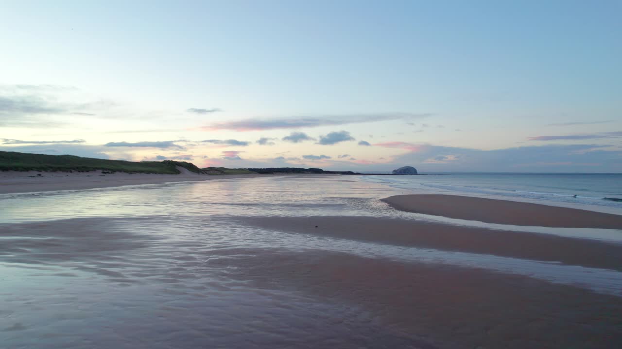 imágenes de drones en movimiento rápido que vuelan cerca de la superficie de una larga playa de arena antes de elevarse por encima de ella hacia una puesta de sol mientras la marea golpea la orilla