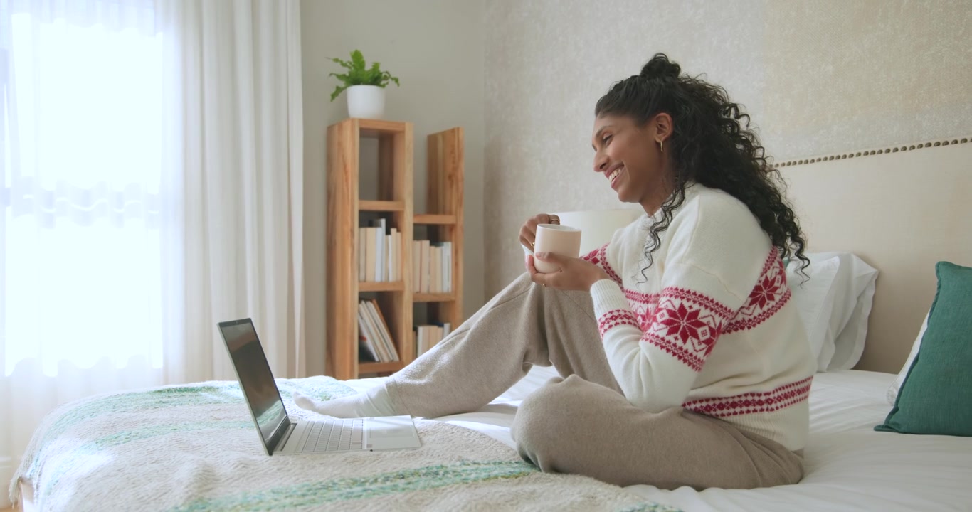 Donna afroamericana con maglione bianco che sorseggia da una tazza, si muove sul letto, sorridendo ai contenuti del laptop