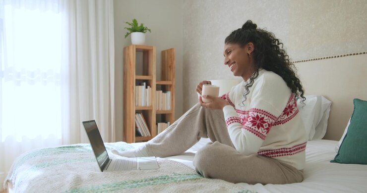 Donna afroamericana con maglione bianco che sorseggia da una tazza, si muove sul letto, sorridendo ai contenuti del laptop